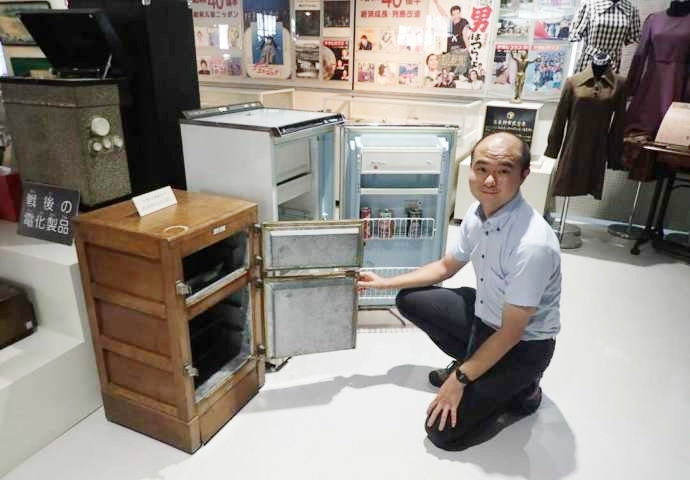 「富良野市博物館」に展示中の氷式冷蔵庫と高度成長期の電気冷蔵庫に並ぶ学芸員の泉さん