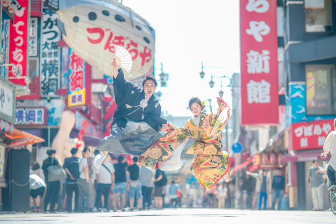 大阪らしい繁華街を背景に和装でジャンプする新郎新婦