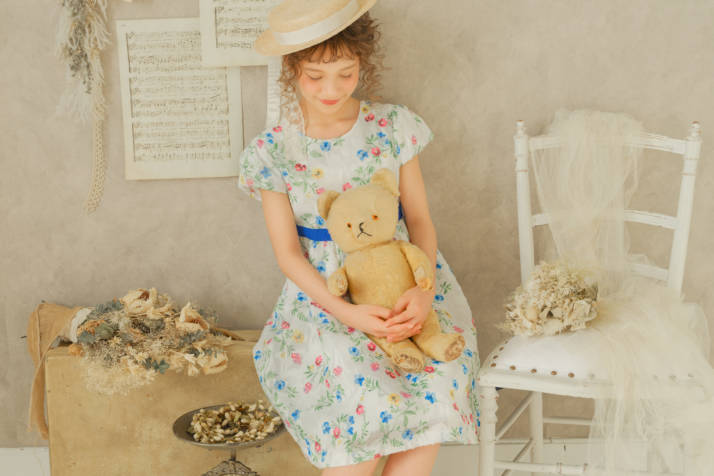 クマのぬいぐるみを膝にのせてほほ笑む花柄ワンピースを着た女の子の写真