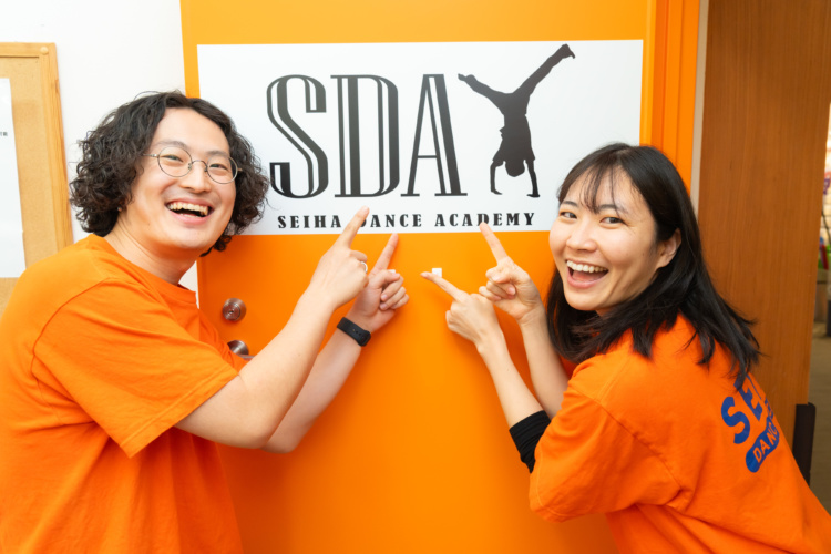 セイハダンスアカデミーの大平さんと齊木さんがスクールのロゴの前でポーズをとるようす
